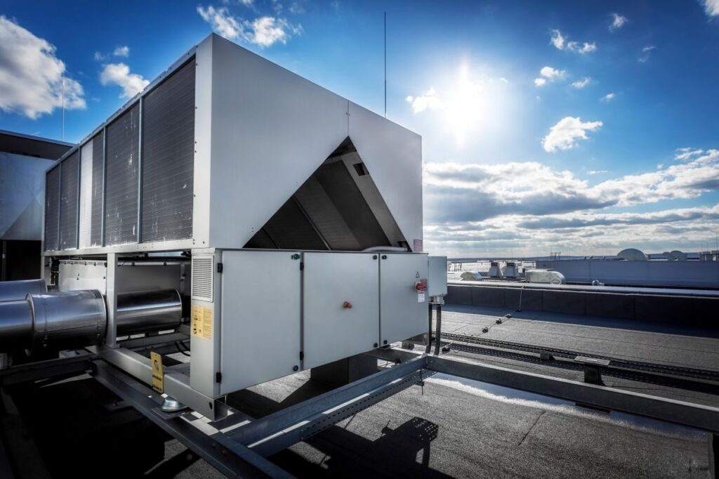 A huge air conditioning unit on the roof of the building. In the background of blue sky with shining sun. Focus is at the front of the air conditioner, the other parts of image slightly blurred. See Less