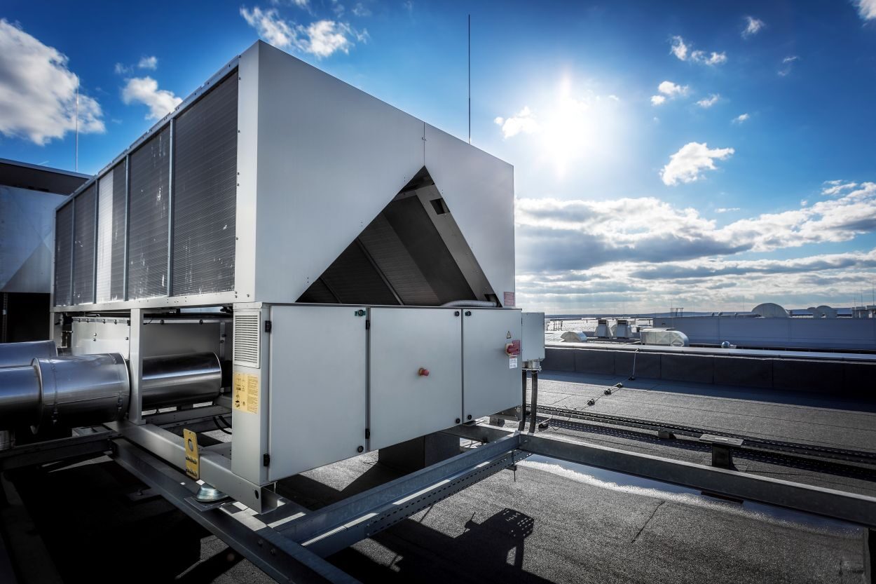 A huge air conditioning unit on the roof of the building. In the background of blue sky with shining sun. Focus is at the front of the air conditioner, the other parts of image slightly blurred. See Less